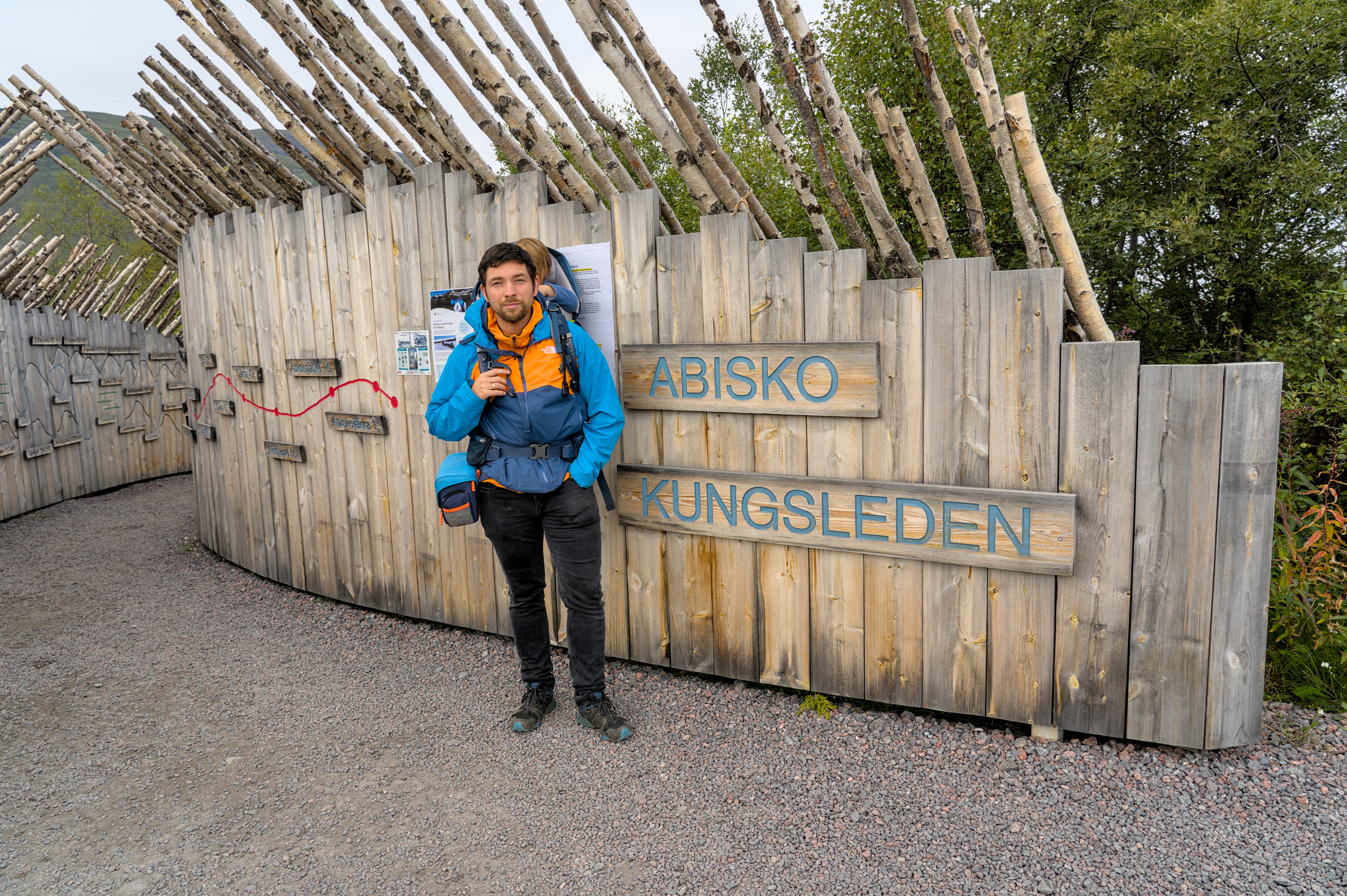 Eine Person mit Wanderkleidung steht vor einem Holzschild am Eingang des Kungsleden in Abisko.