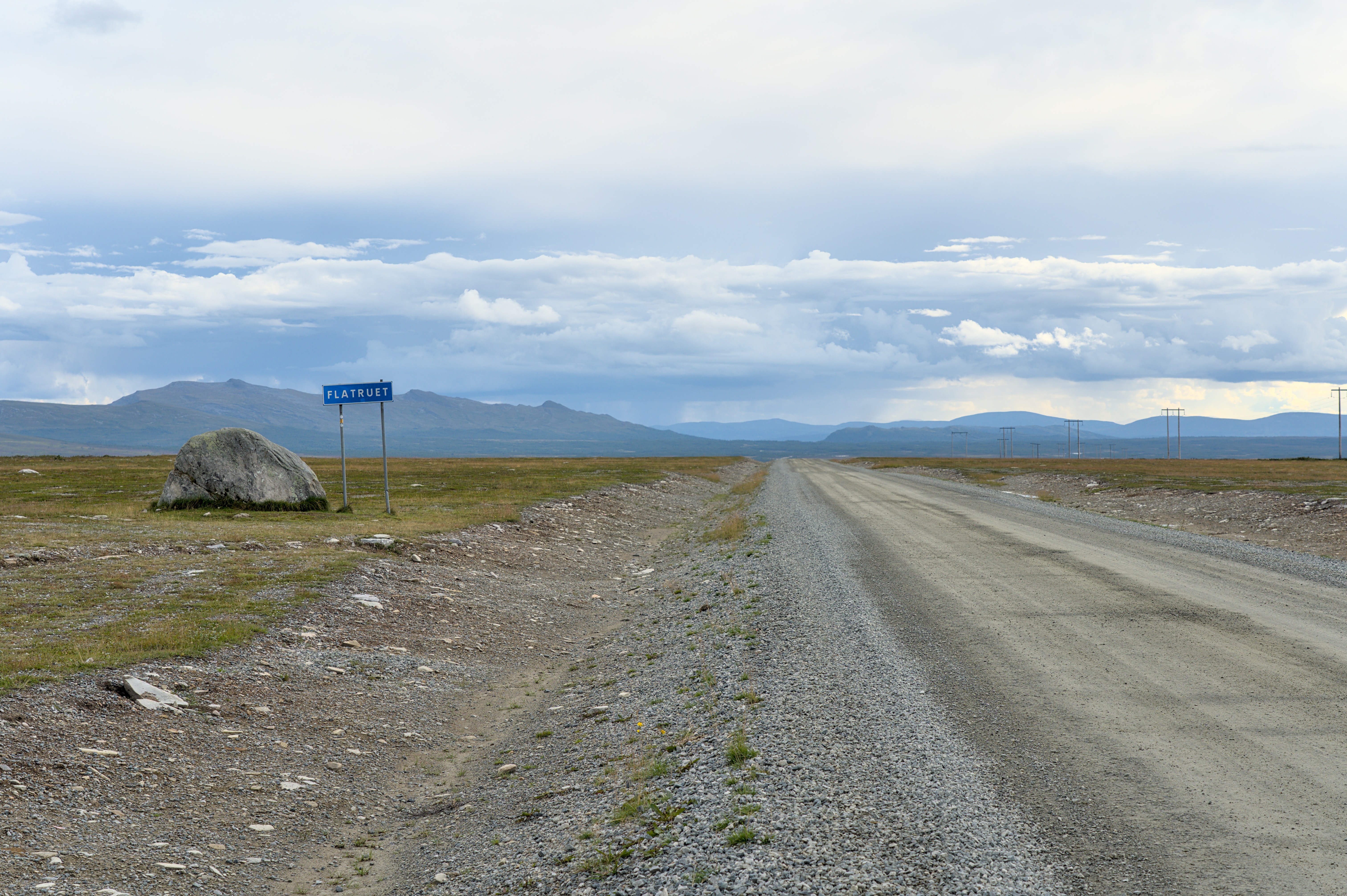 Weitläufige Landschaft mit Schotterstraße auf dem Flatruet, einem Gebirgspass in Schweden, und einem markanten Felsblock neben einem Straßenschild.
