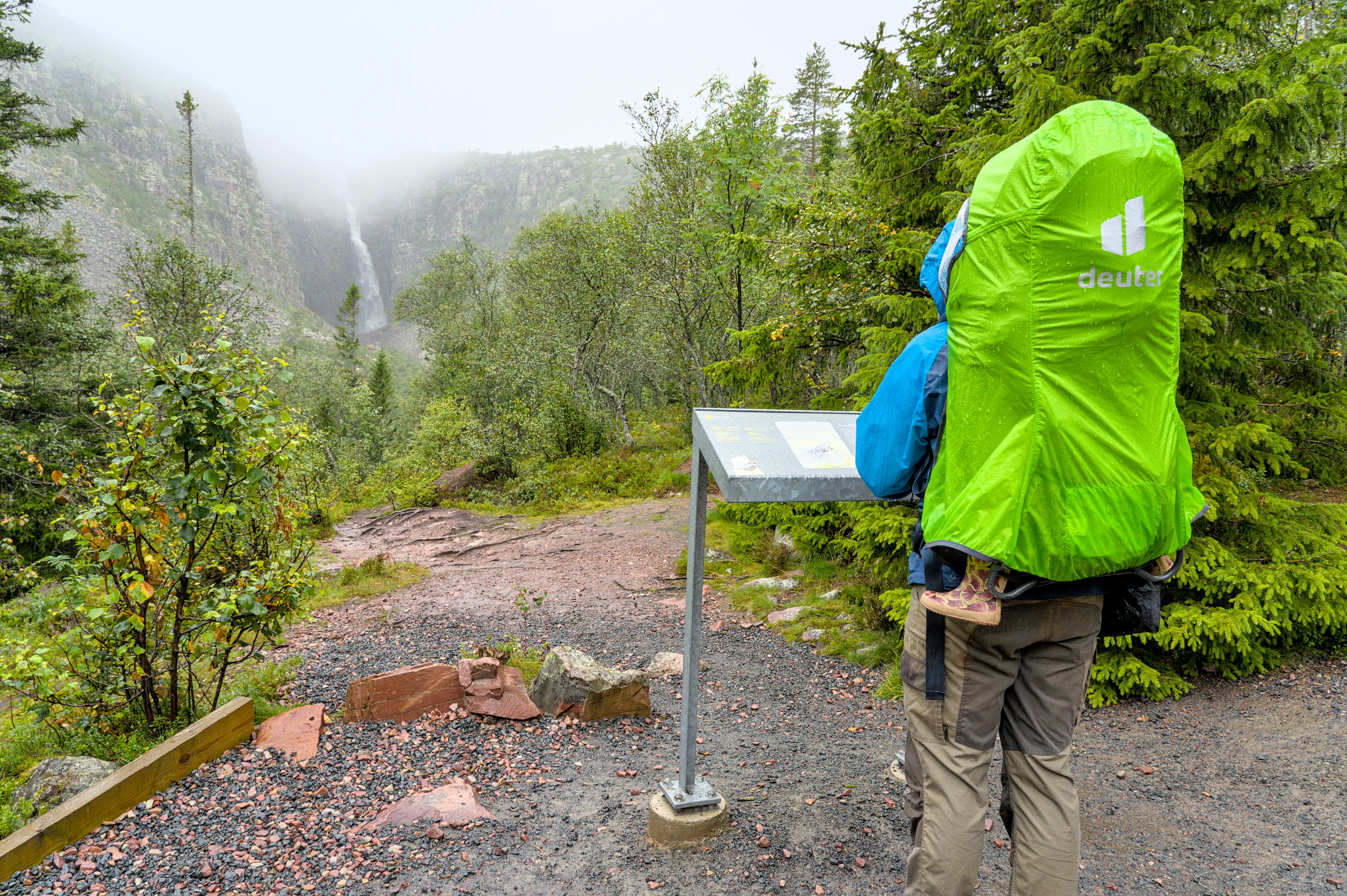 Ein Wanderer mit einem grünen Rucksack-Cover steht vor einer Informationstafel im Skuleskogen Nationalpark bei nebligem Wetter. Im Hintergrund ist ein Wasserfall zu sehen.