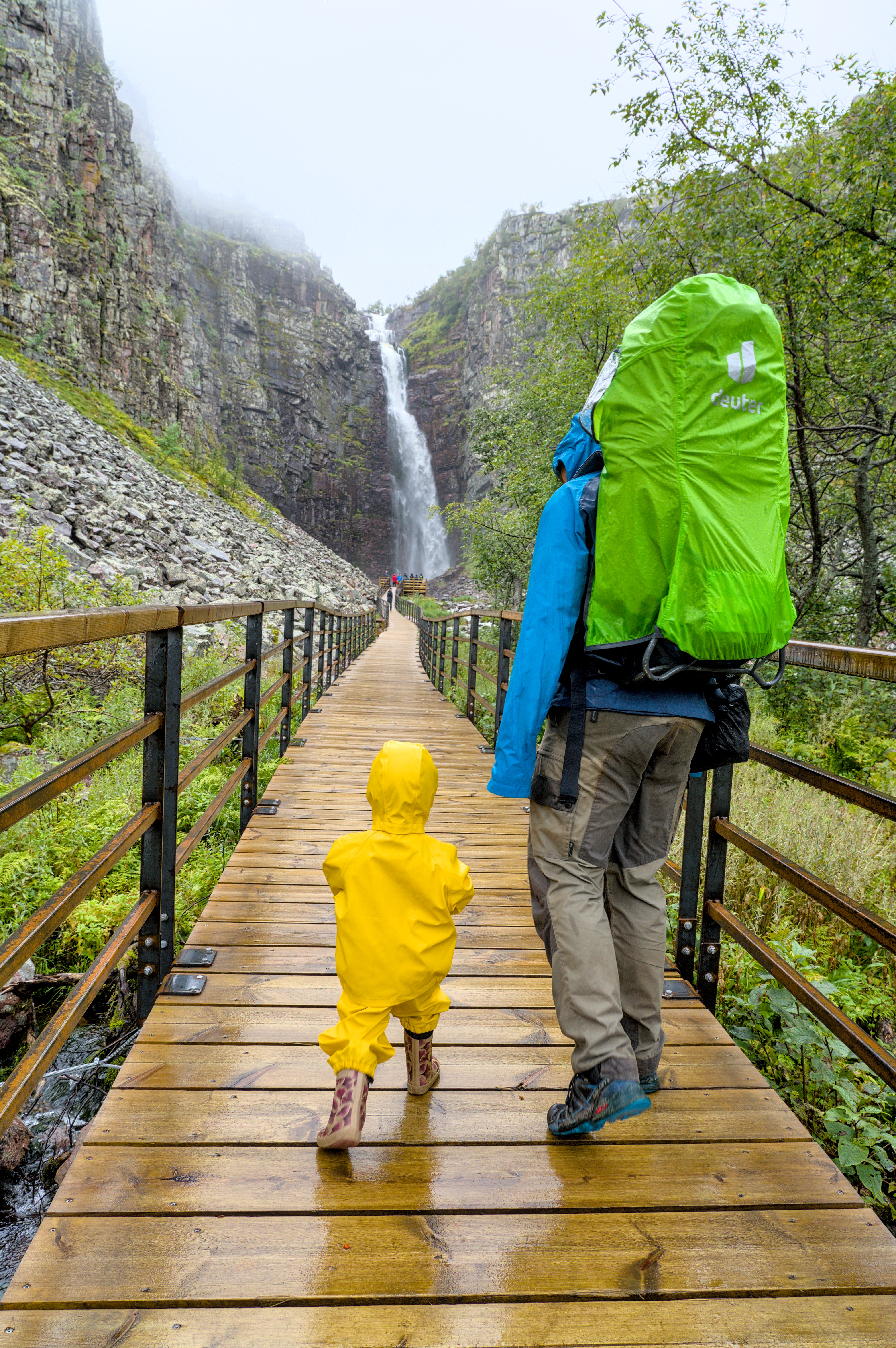 Vater und Kind in Regenkleidung auf einem Holzweg vor dem Njupeskär-Wasserfall in Schweden.